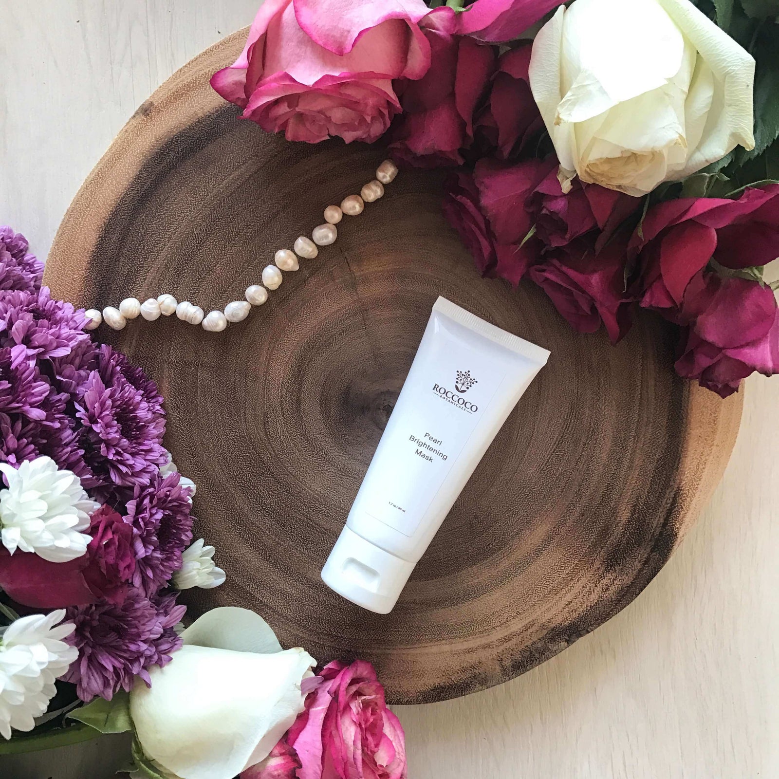 white pearl mask tube on round wood block with string of fresh pearl next to it surrounded by white, pink and purple roses